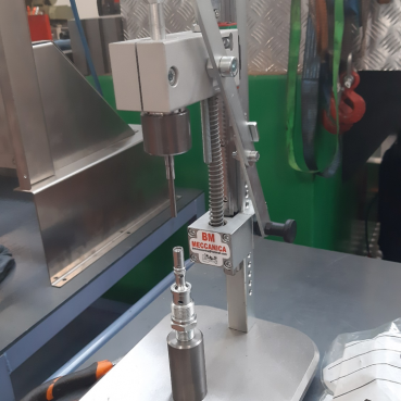 Un trapano a colonna da banco &egrave; montato su un banco da lavoro. La macchina ha una base metallica su cui viene fissata una parte cilindrica. Sullo sfondo si possono vedere vari strumenti e attrezzature personalizzate, tra cui un cacciavite e una serie di anelli appesi ai ganci. Sulla macchina &egrave; applicata un'etichetta "BM MECCANICA".