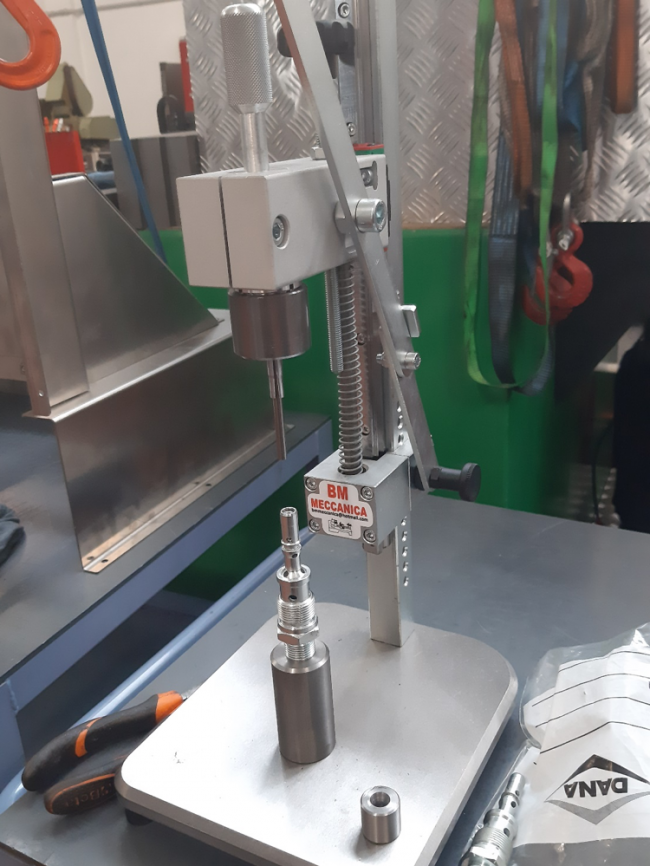 Un trapano a colonna da banco è montato su un banco da lavoro. La macchina ha una base metallica su cui viene fissata una parte cilindrica. Sullo sfondo si possono vedere vari strumenti e attrezzature personalizzate, tra cui un cacciavite e una serie di anelli appesi ai ganci. Sulla macchina è applicata un'etichetta "BM MECCANICA".