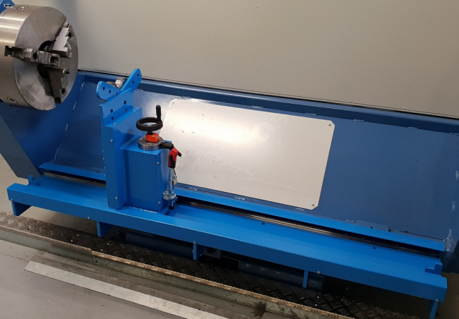 Contro una parete liscia si trova una macchina industriale blu con base in metallo e vari componenti, tra cui una maniglia e delle manopole. Per la lavorazione di parti metalliche sembrano essere utilizzate le attrezzature personalizzate, dotate di mandrino e pannelli di protezione. I dintorni sono puliti e ben tenuti.