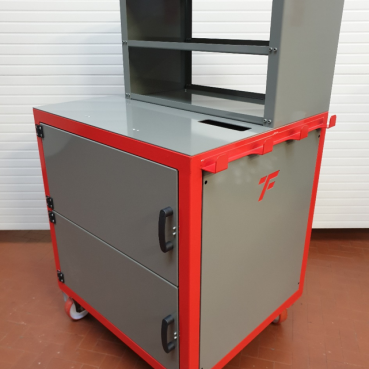 Una postazione di lavoro su ruote in metallo grigio e rosso, dotata di due cassetti inferiori, un ampio vano e due ripiani aperti nella parte superiore. L'unit&agrave; ha maniglie nere, un logo con la lettera "F" sul lato e incorpora attrezzature personalizzate. &Egrave; posizionato contro una parete bianca a coste su un pavimento piastrellato marrone.