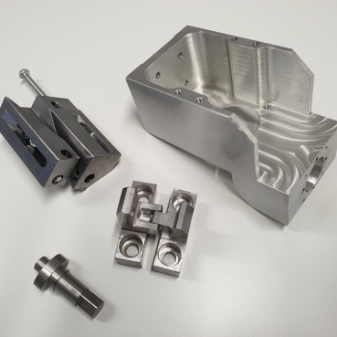 Un insieme di parti metalliche lavorate disposte su una superficie bianca. I componenti dell'attrezzatura personalizzata includono un pezzo grande, parzialmente chiuso con tagli intricati, pi&ugrave; pezzi rettangolari e cilindrici pi&ugrave; piccoli con fori precisi e un lungo bullone con una rondella.