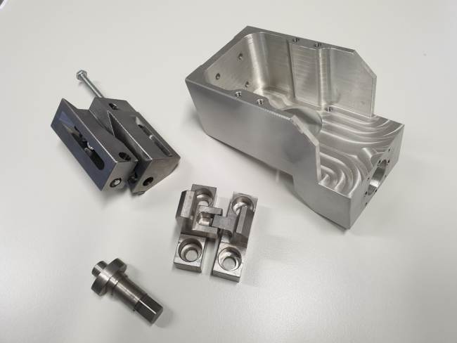 Un insieme di parti metalliche lavorate disposte su una superficie bianca. I componenti dell'attrezzatura personalizzata includono un pezzo grande, parzialmente chiuso con tagli intricati, più pezzi rettangolari e cilindrici più piccoli con fori precisi e un lungo bullone con una rondella.