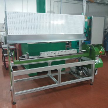 Immagine di una macchina industriale in un'officina. La macchina ha un grande tabellone metallico forato e poggia su un telaio metallico con diversi componenti e controlli. Lo sfondo del laboratorio presenta strumenti, attrezzature, attrezzature personalizzate e macchinari vari su un pavimento di piastrelle marroni.