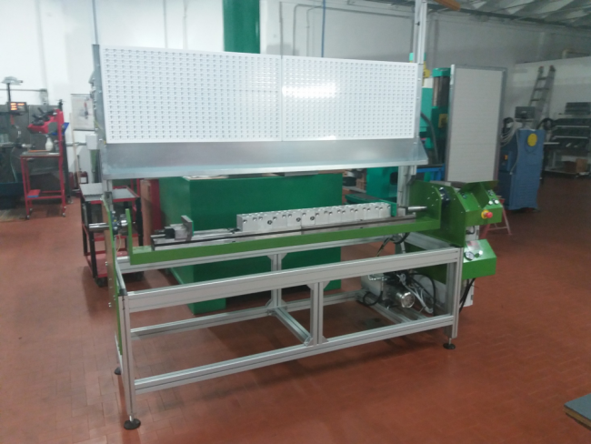 Immagine di una macchina industriale in un'officina. La macchina ha un grande tabellone metallico forato e poggia su un telaio metallico con diversi componenti e controlli. Lo sfondo del laboratorio presenta strumenti, attrezzature, attrezzature personalizzate e macchinari vari su un pavimento di piastrelle marroni.